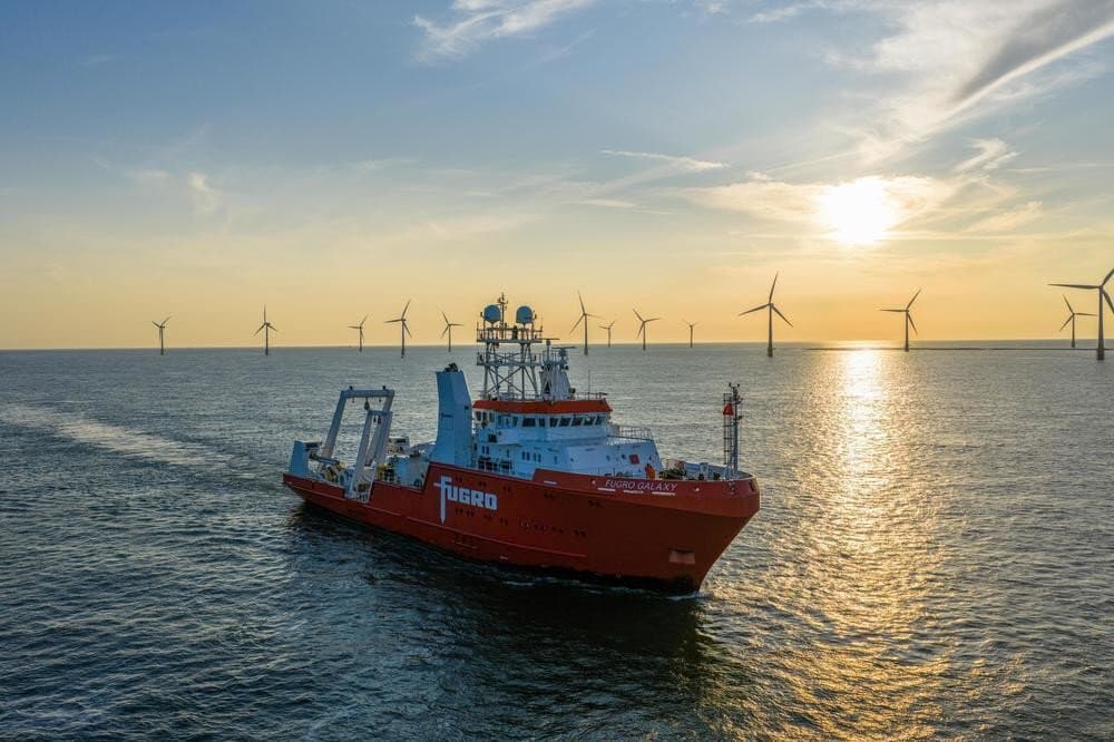 Fugro Galaxy offshore Great Yarmouth in UK, sailing at sunset in open sea, with multiple wind turbines visible on the horizon and sunlight reflecting across the water.